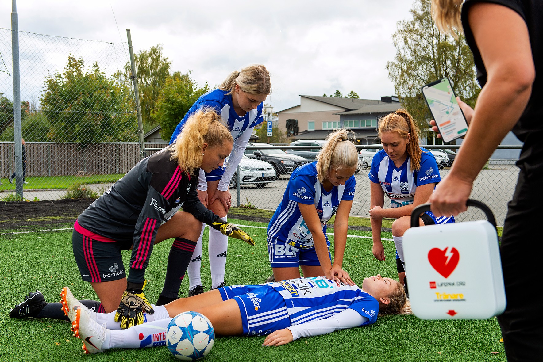 En flicka har fått hjärtstopp under en fotbollsmatch. Kamraterna utför HLR medan en SMS-Livräddare anländer med hjärtstartare.